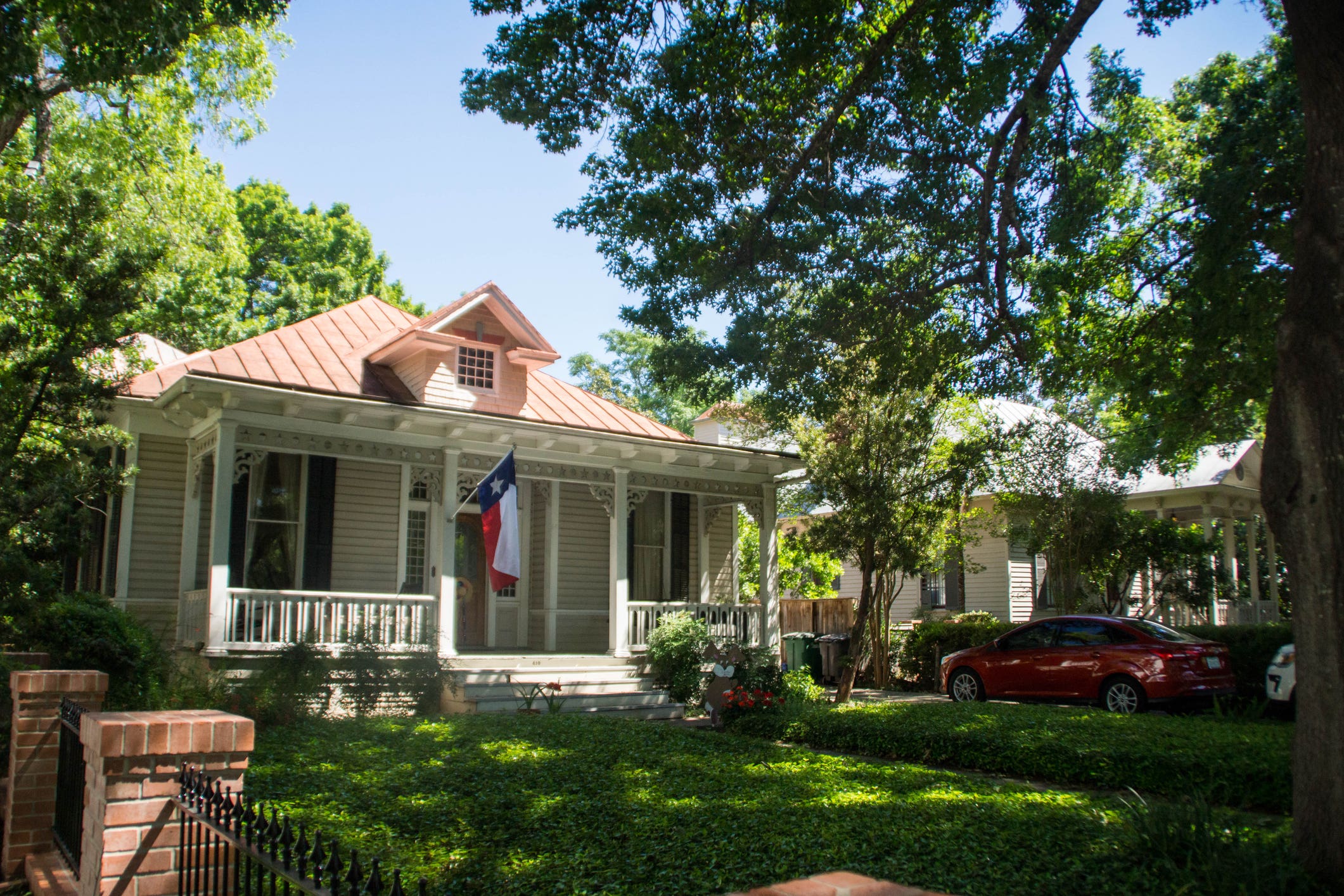 Single-family home with Texas flag in fenced yard with lots of trees and red sedan in driveway.