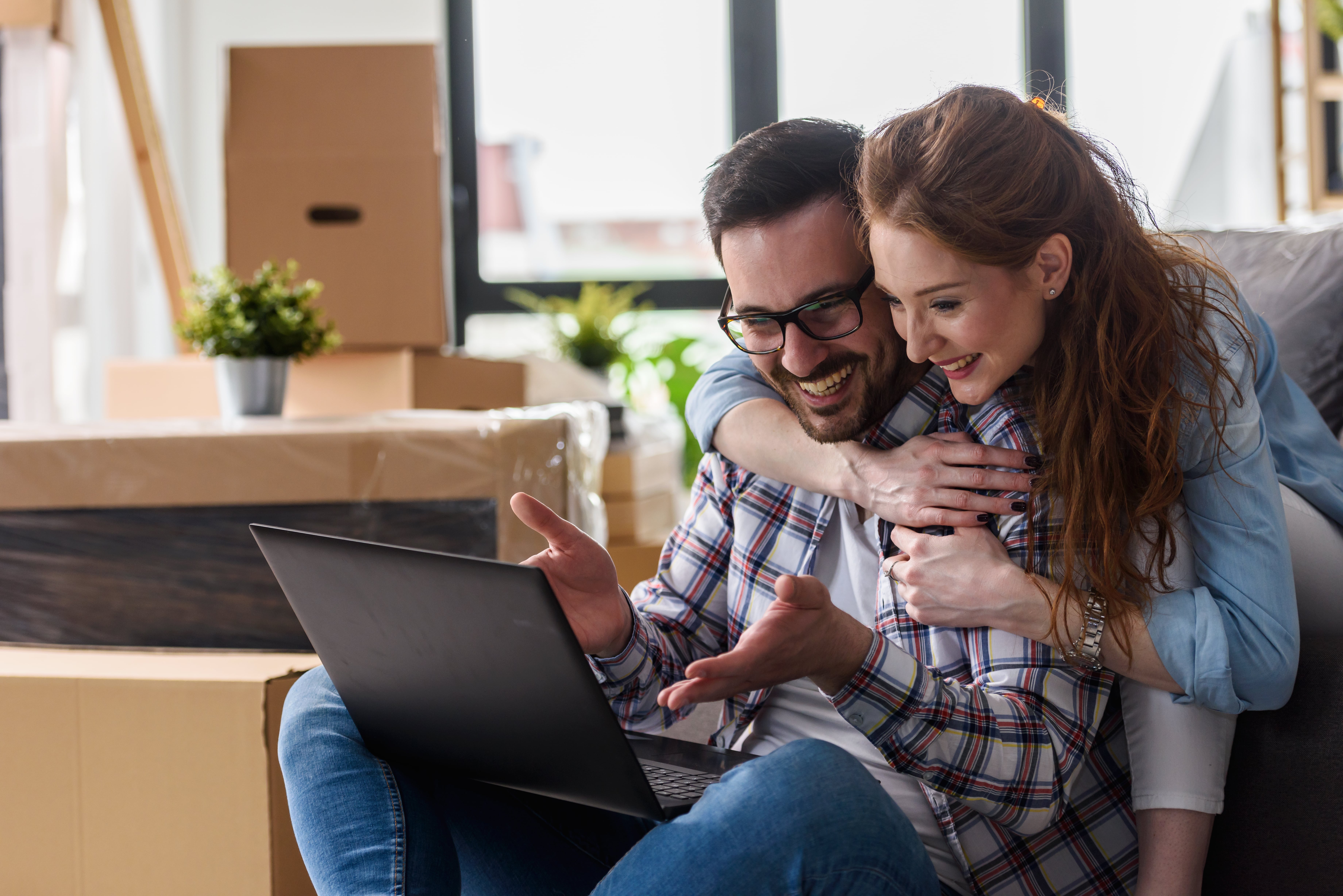 Closeup of a young couple sitting on the floor of their new home, surrounded by boxes, applying for insurance on a laptop