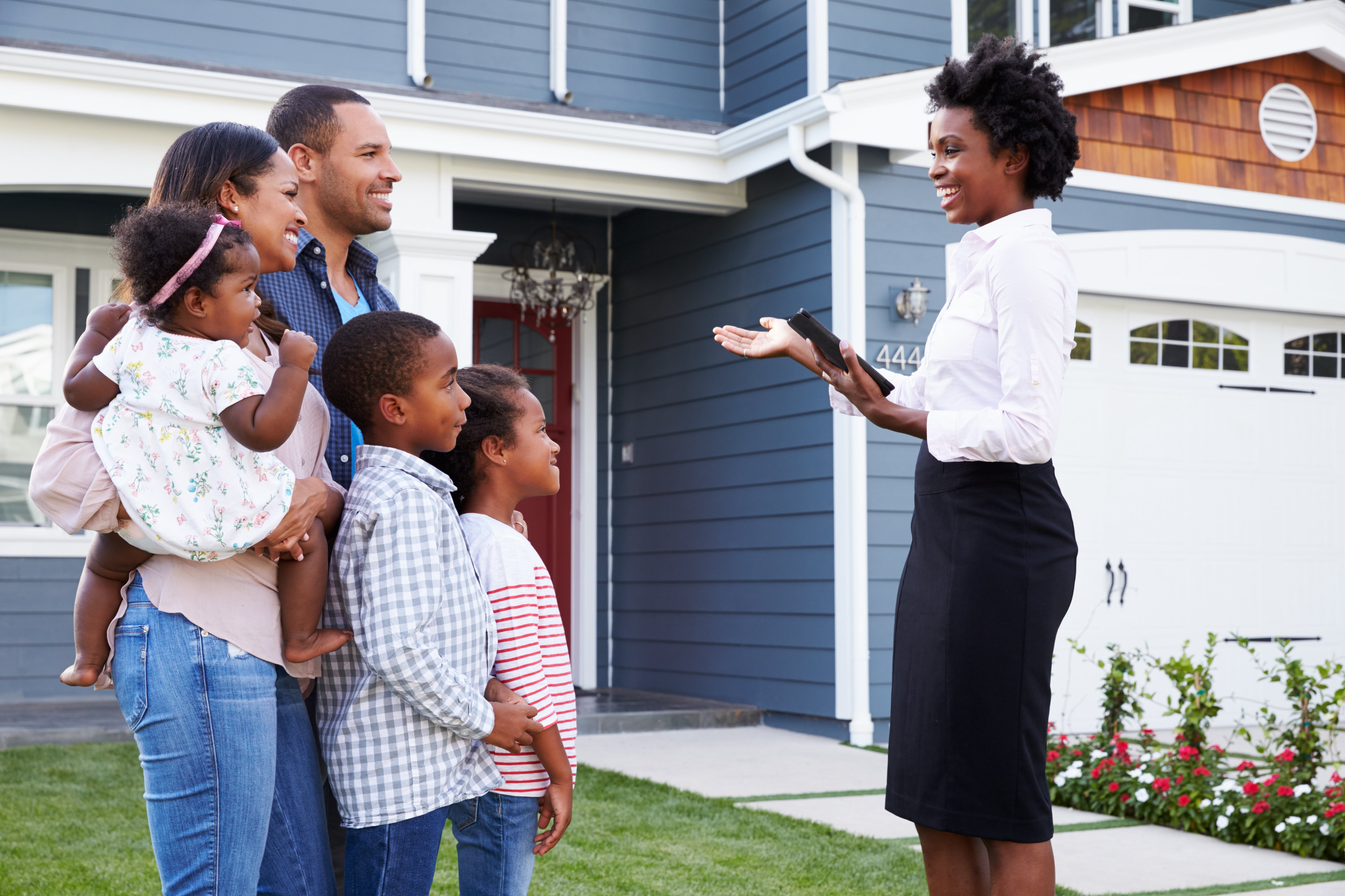 A female real estate agents meets with a couple and their three children in front of a blue house.