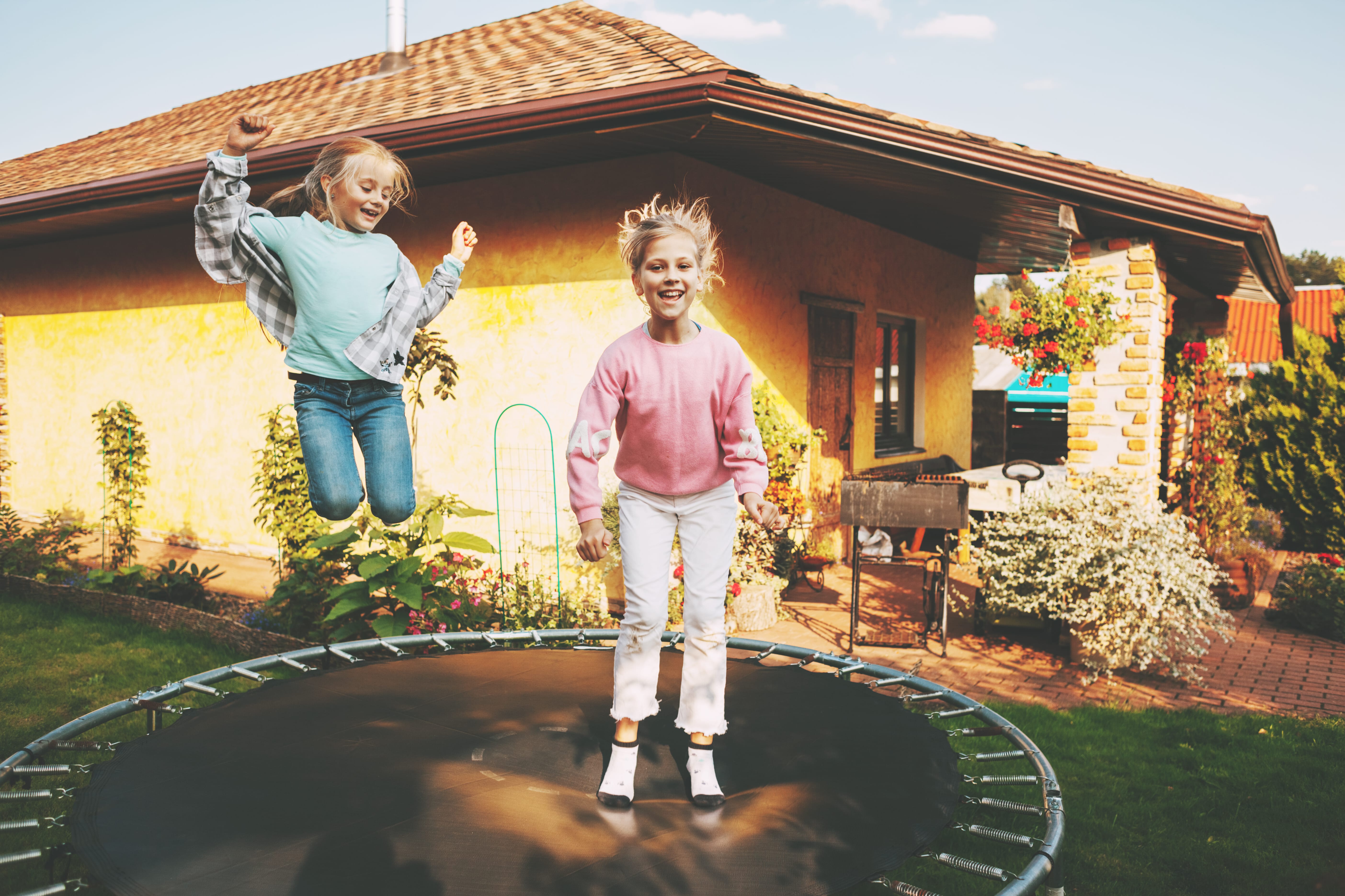 two young girls jumping on a trampoline in front of a one-story home
