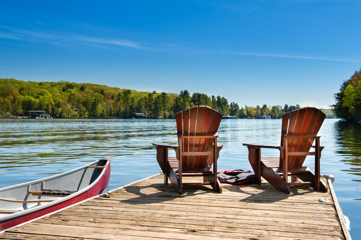 Two chairs on a wooden dock overlooking the blue water of a lake; a red canoe is tied to the pier and life jackets are visible near the chairs
