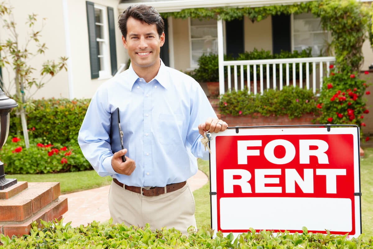 A landlord stands by a For Rent sign in front of a home