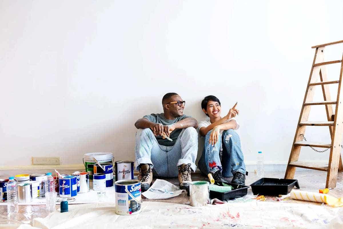 A young couple sit on the floor surrounded by paint cans and a ladder