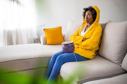 Worried young woman in raincoat holding bucket under water drops.
