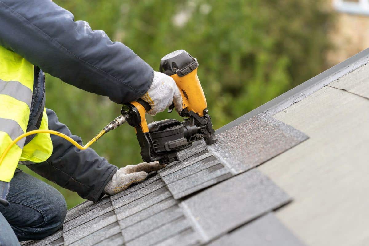 A roofer in uniform using a pneumatic nail gun to rplace asphalt shingles on a home's roof