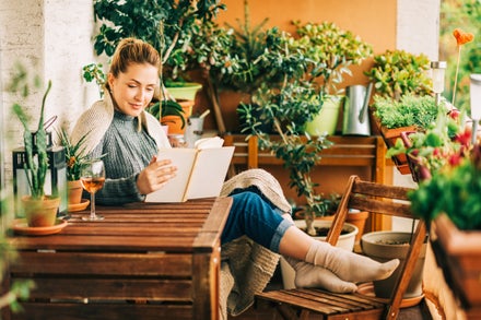 a woman sits at a garden table with a glass of wine, reading a book