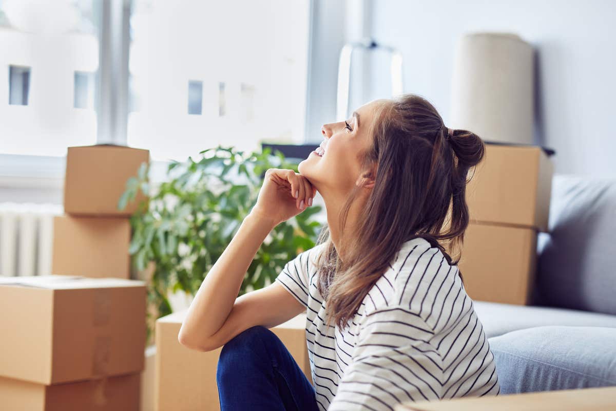 A smiling woman sits in her new home surrounded by moving boxes