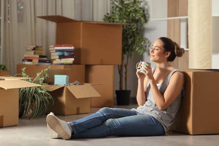 A young woman sits on the floor, surrounded by moving boxes, drinking coffee