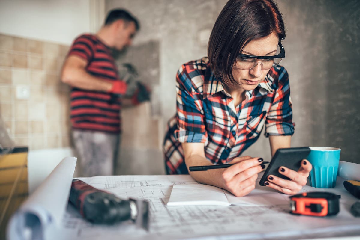 a woman wearing safety glasses works on a calculator while a man behind her takes tile off a wall
