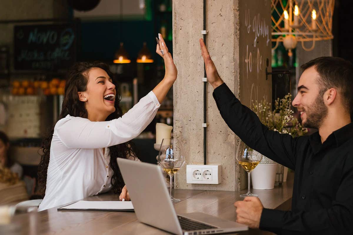 A young couple sits in a restaurant with a laptop and high-five each other
