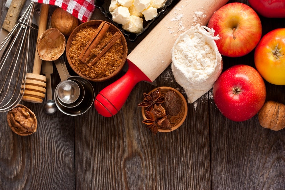 Overhead shot of baker's wooden table with apples, flour, cinnamon, and walnuts, plus a whisk, rolling pin, and measuring spoons