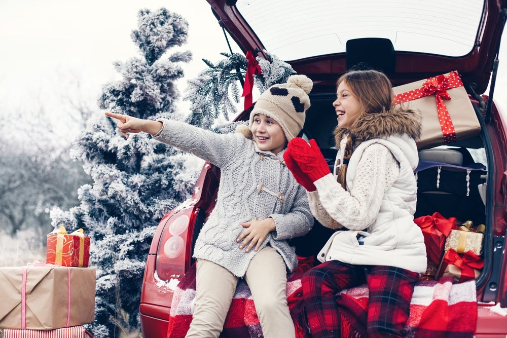 Image of two young girls dressed for winter sitting in the back of a hatchback loaded up with holiday presents