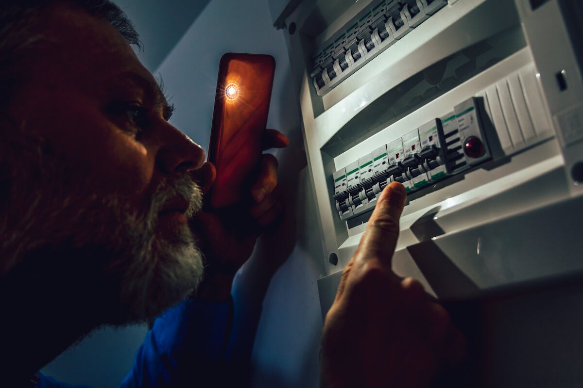 Man in complete darkness holds a phone to investigate a home fuse box during a power outage 