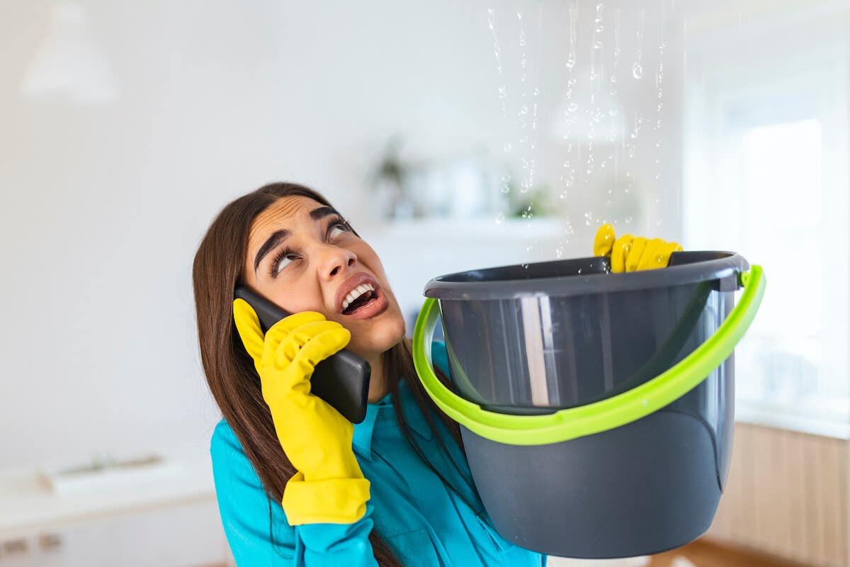 Young woman calls a plumber while water leaks from her ceiling into a bucket.