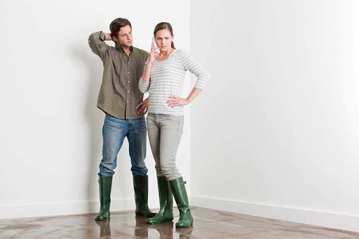 A young couple stand on a flooded floor wearing rainboots while one calls their insurer