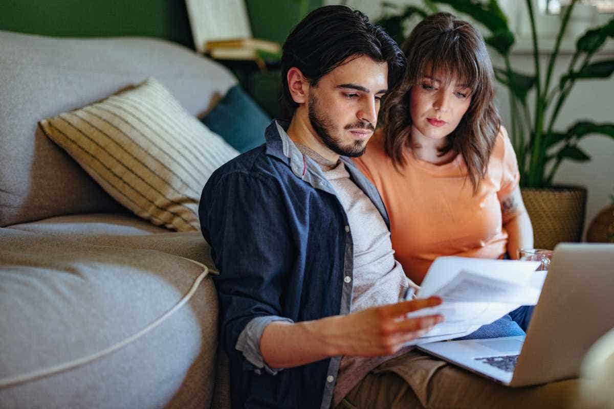 A serious-looking couple holding bills and doing home finances together on a laptop computer in the living room