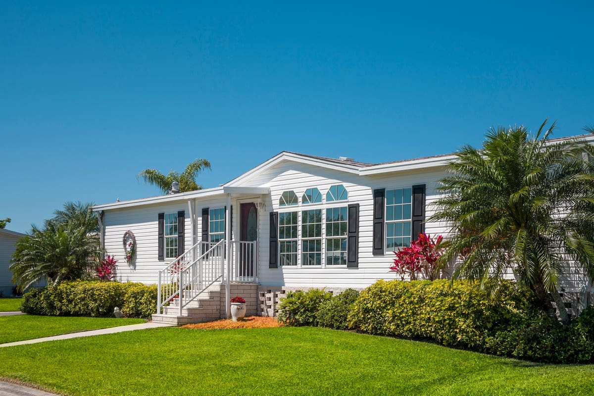 white manufactured home with brown shutters and palm trees on the front lawn and a blue sky overhead