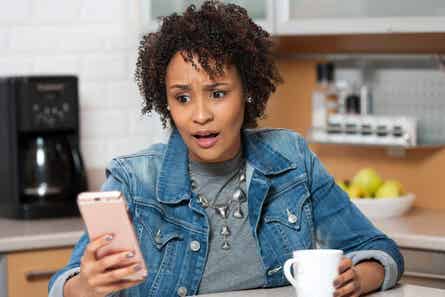A young woman looks at her phone with shock while sitting in kitchen