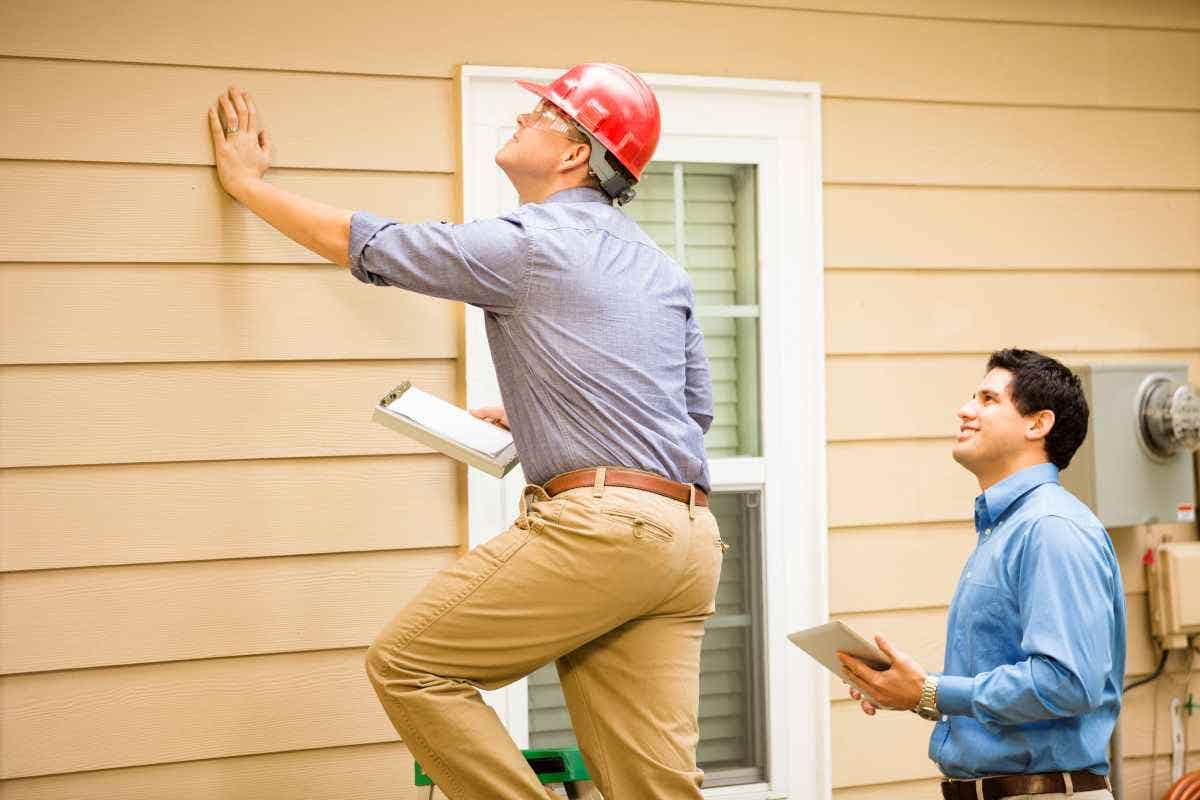 A claims adjuster stands on a ladder outside of a home while the homeowner looks on