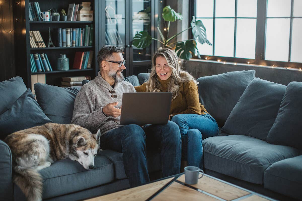 man and woman and dog on a blue couch looking at their laptop and smiling