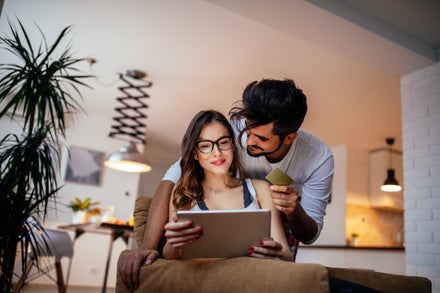 A young couple sit on their couch; the woman holds a tablet and the man holds a credit card
