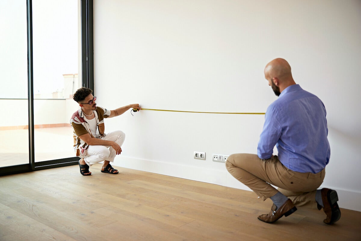 Two men couch by a white wall with a measuring tape stretched between them