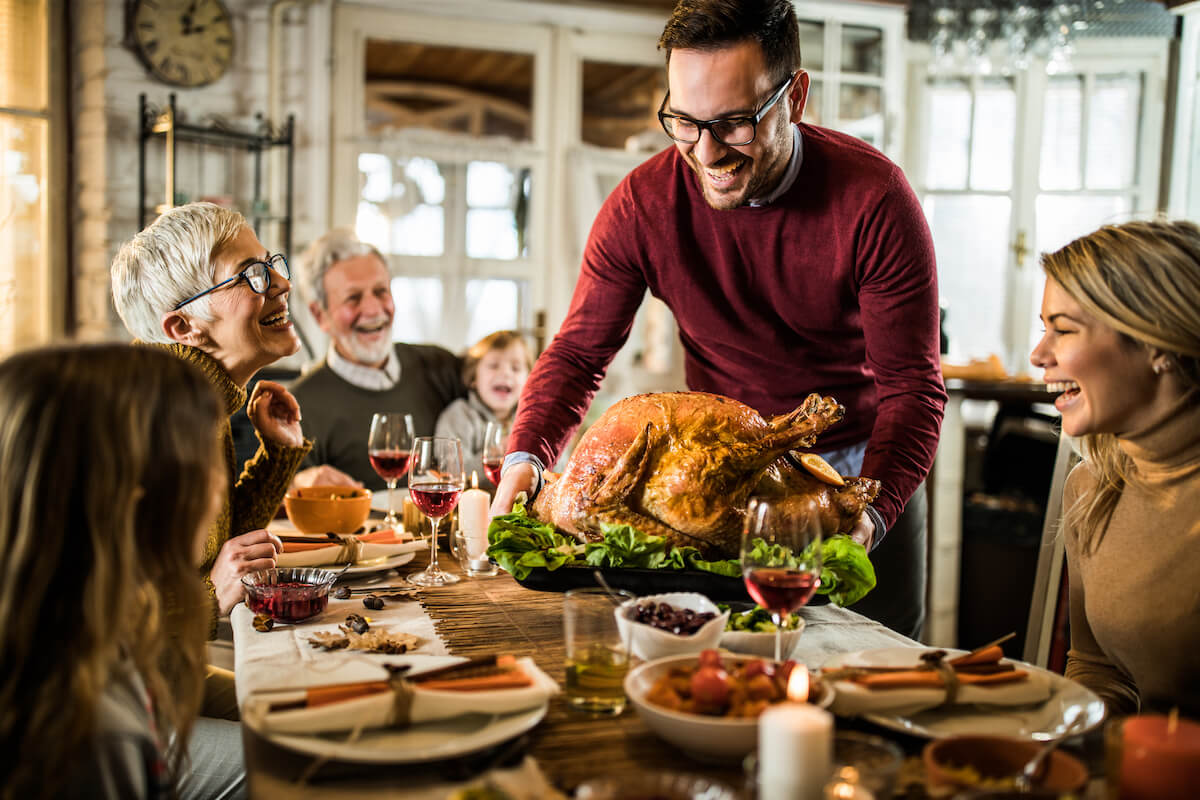 smiling family gathered around a large wooden dinner table while a man in glasses and a red sweater serves a turkey