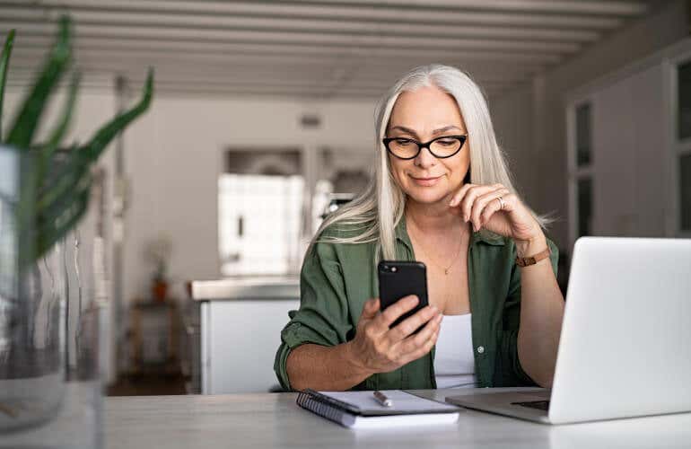 Woman with glasses and gray hair looks at her phone while sitting in front of a laptop