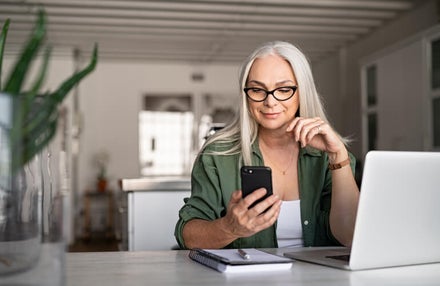 A woman sits at her kitchen table with a laptop and paper notebook, looking at her phone